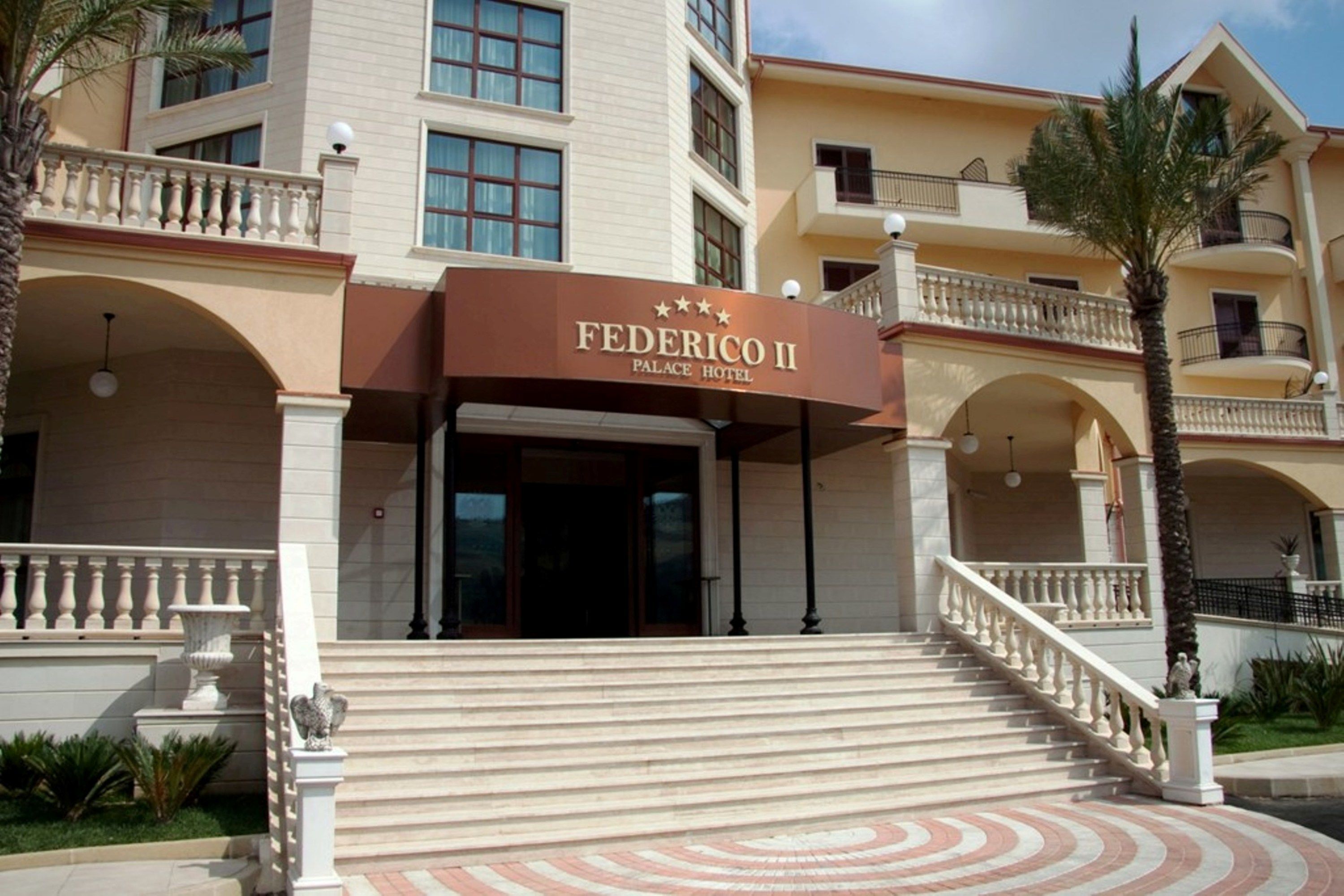 The exterior of the Federico II Palace Hotel with a grand entrance, multiple balconies, and a well-maintained garden area.