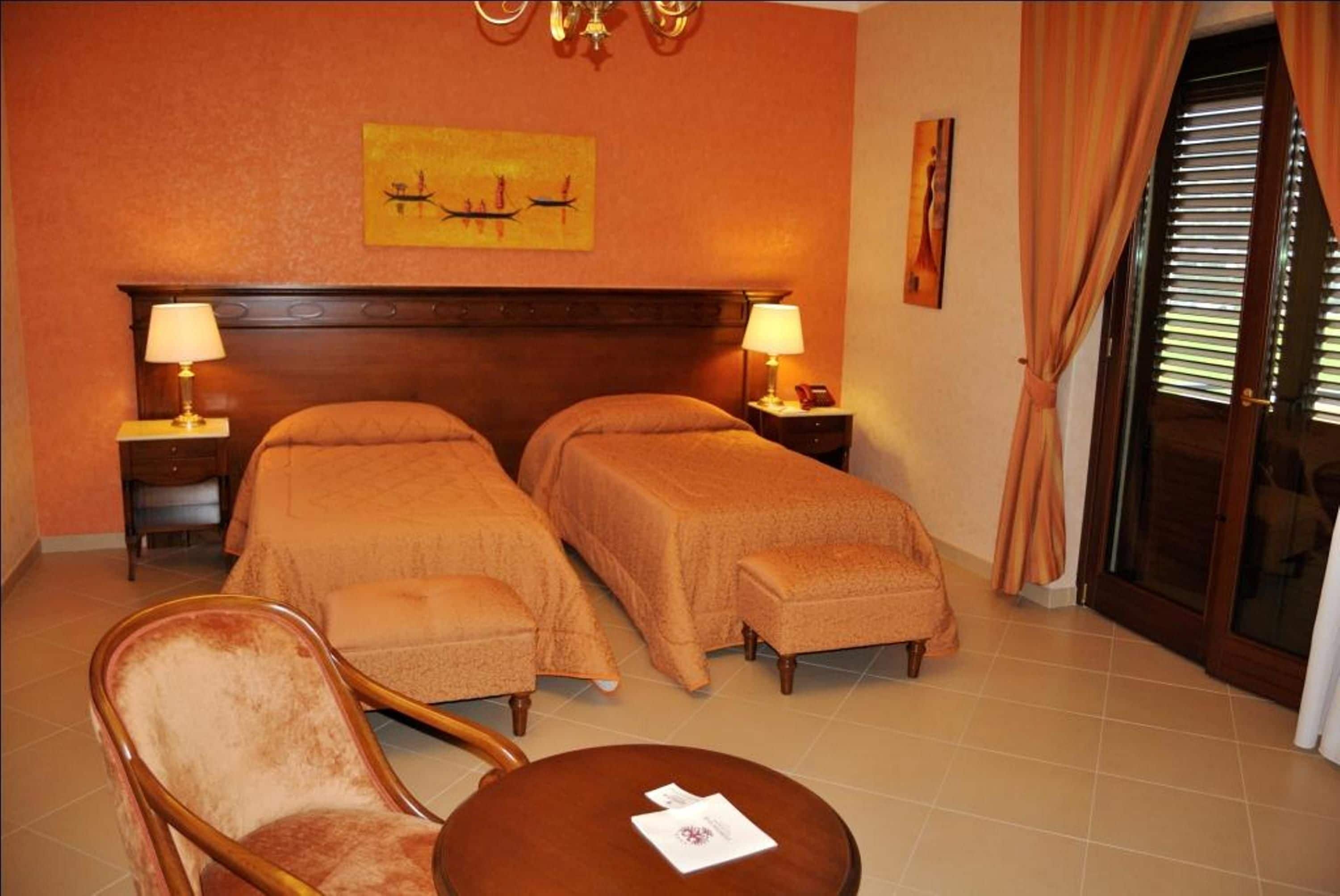 Habitación de hotel con dos camas individuales, una silla de madera, una mesita y vistas al exterior a través de un ventanal.