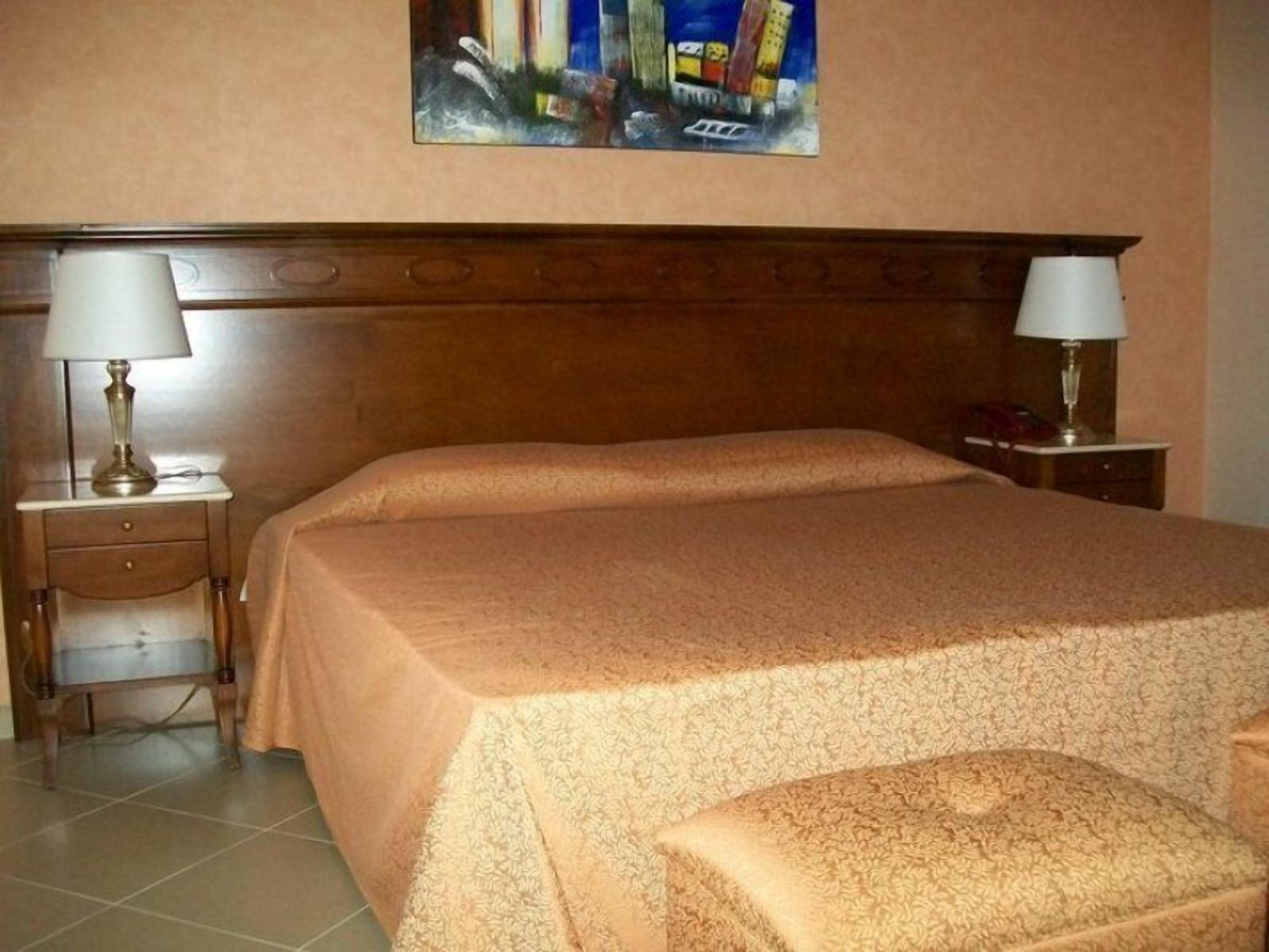 A neatly made bed with a patterned bedspread, two bedside tables with lamps, a small drawer, and a painting on the wall.