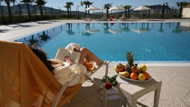 Zona junto a la piscina con una persona relajándose en una tumbona, una mesa con frutas y sombrillas al fondo.