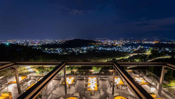 An outdoor dining area with tables and chairs, overlooking a cityscape at night.