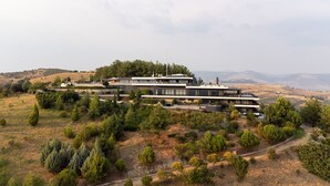 A modern multi-story building with a flat roof, surrounded by a landscaped garden and trees, set against a hilly backdrop.