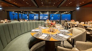 A restaurant with a round table set for dining, a green upholstered booth, and a view of the cityscape through large windows.
