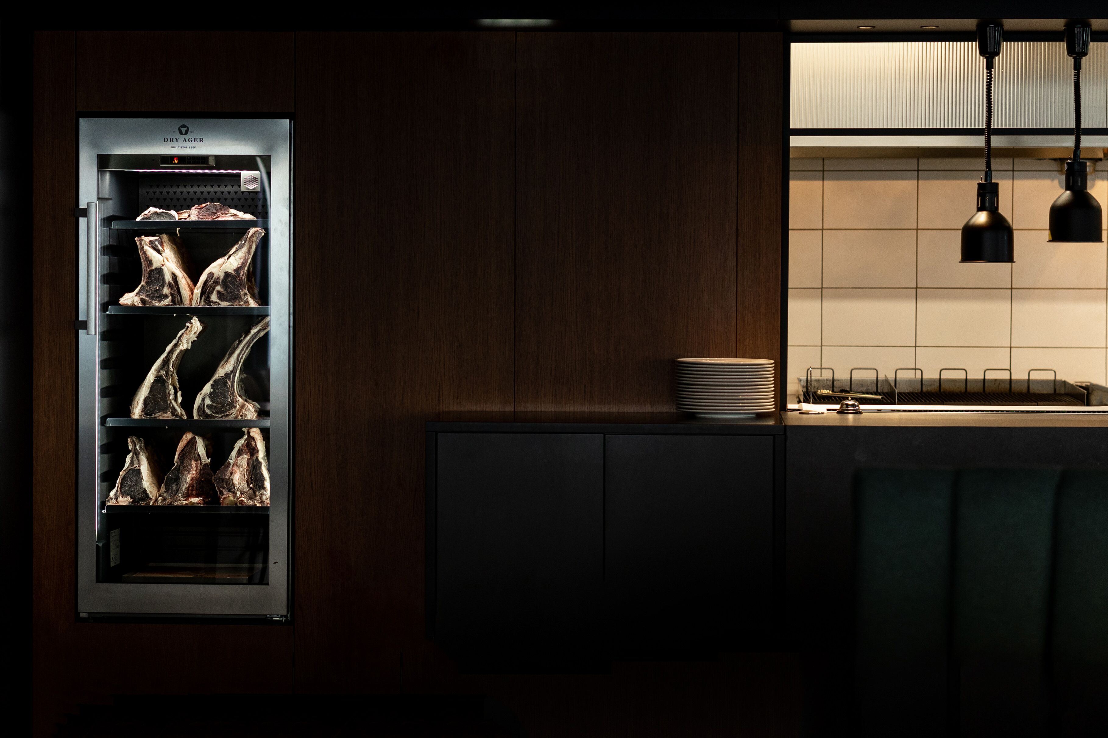 A kitchen with a built-in refrigerator containing several steaks, a dark wooden cabinet, and a stack of plates on the counter.