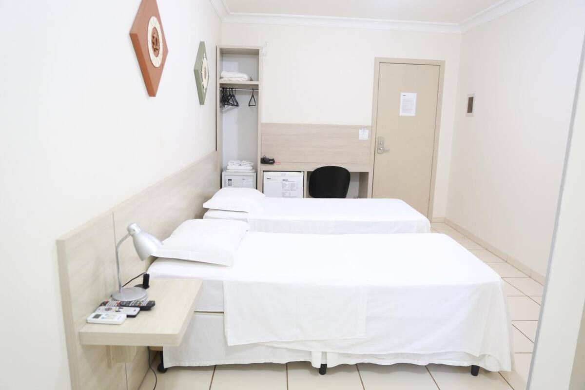 A hotel room with two single beds, a desk with a lamp, a chair, a wardrobe, and a small desk with a computer.