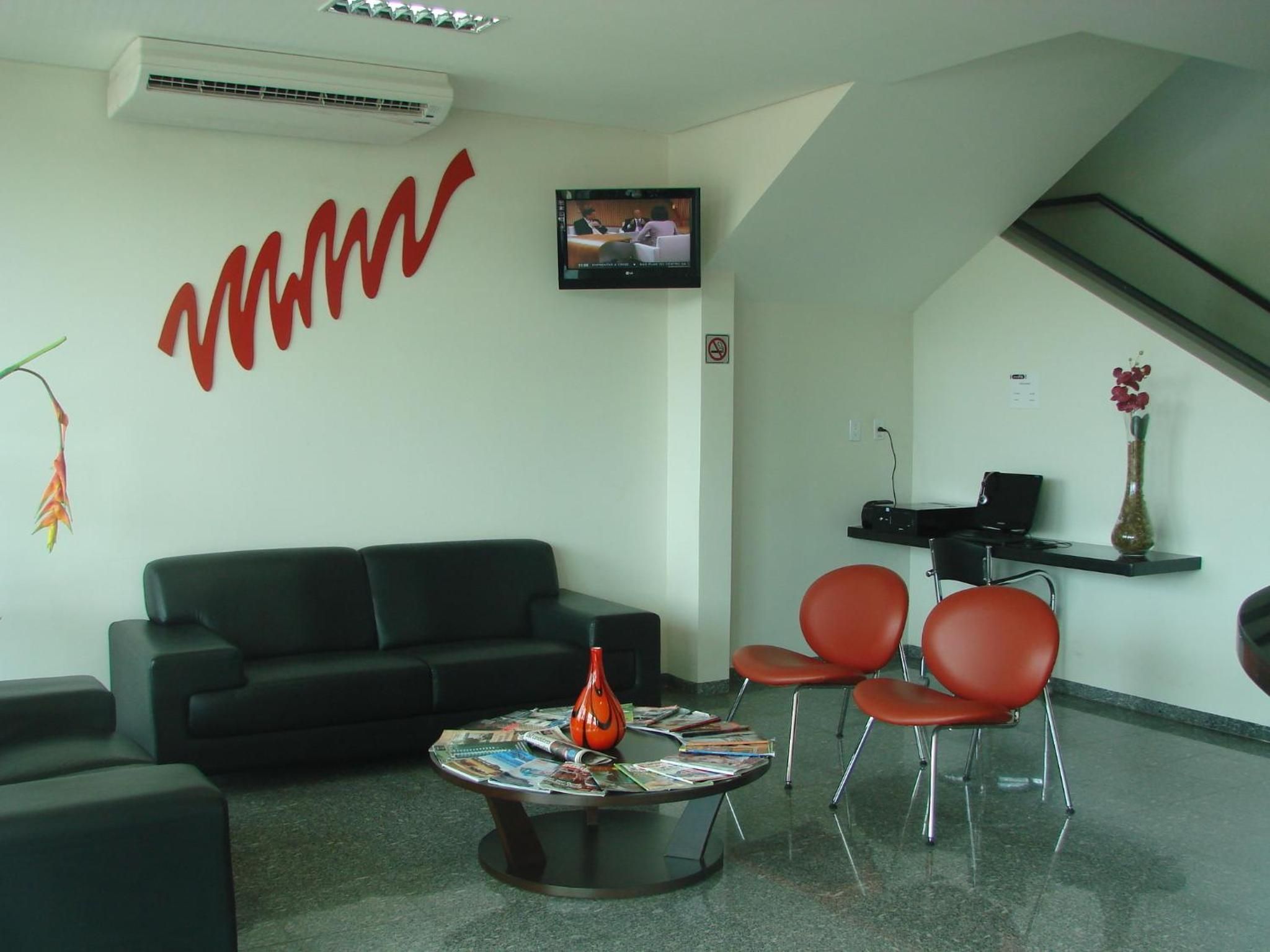 Una habitación con un sofá negro, una mesa redonda con revistas, sillas rojas, un televisor en la pared y un cartel de prohibido fumar.
