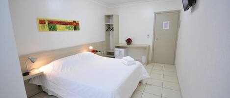 Habitación de hotel con cama, mesita de noche, lámpara, y un pequeño escritorio con espejo y un jarrón de flores.