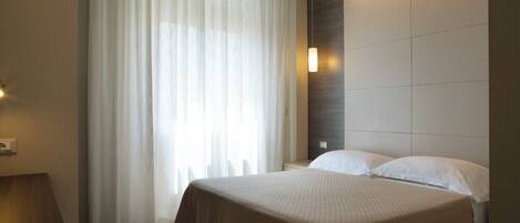 Quarto de hotel com cama, mesa de cabeceira, janela com cortinas leves e luminária na parede.
