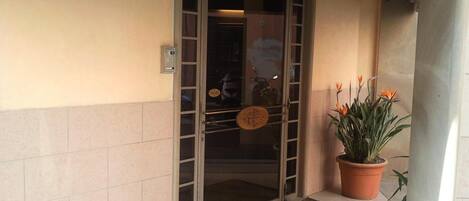 Entrada de hotel con puerta de vidrio, teclado de seguridad y planta con flores anaranjadas.