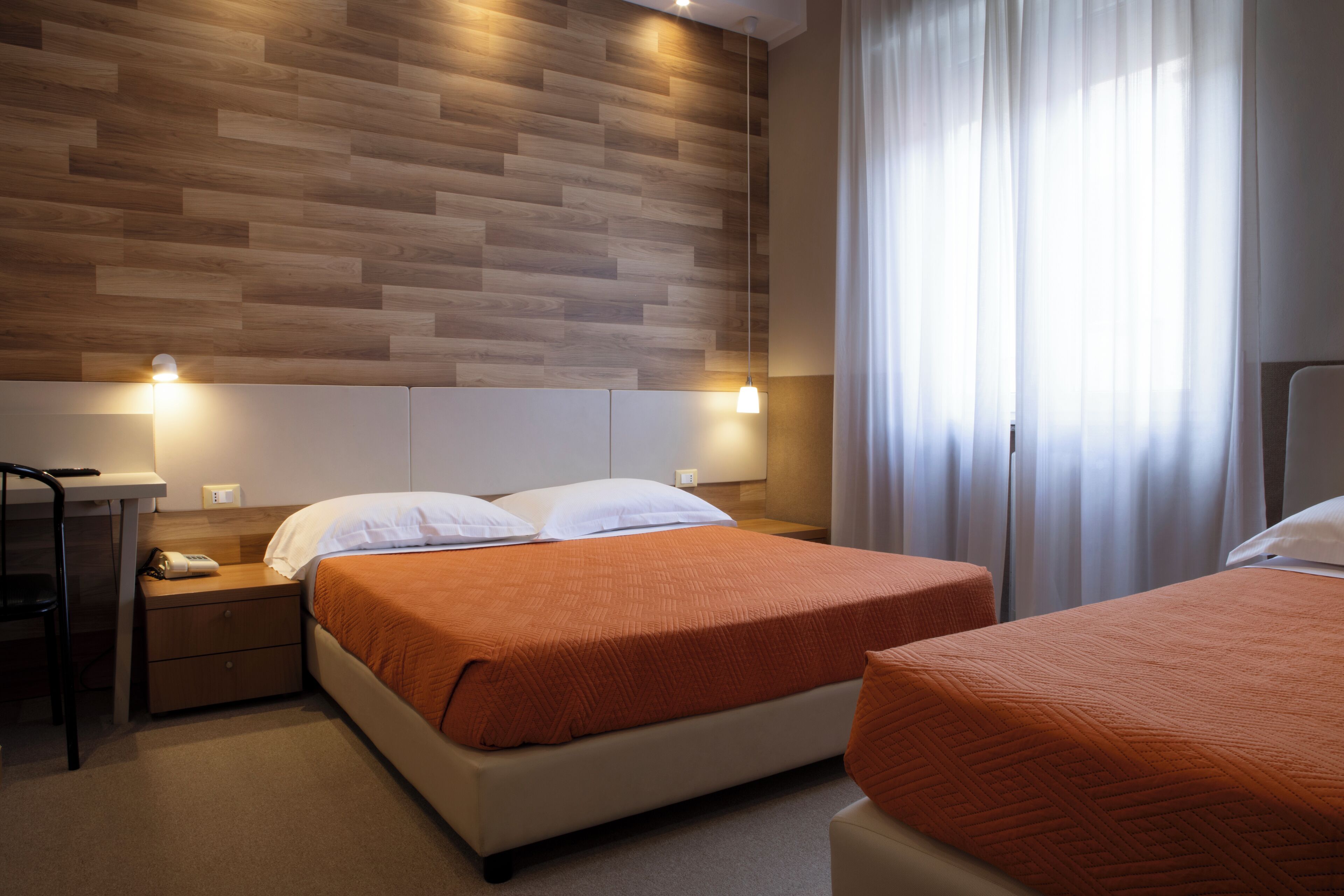 A hotel room with a wooden headboard, a bed with an orange bedspread, a bedside table with a lamp, and a window with curtains.