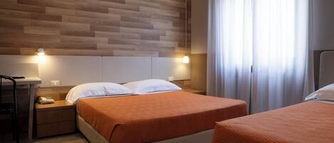 A hotel room with a wooden headboard, a bed with an orange bedspread, a bedside table with a lamp, and a window with curtains.