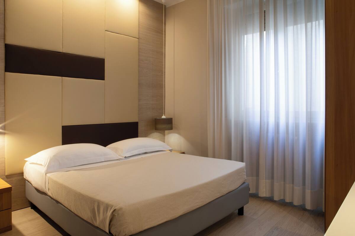 A modern hotel room with a large bed, a bedside table, a lamp, and sheer curtains.