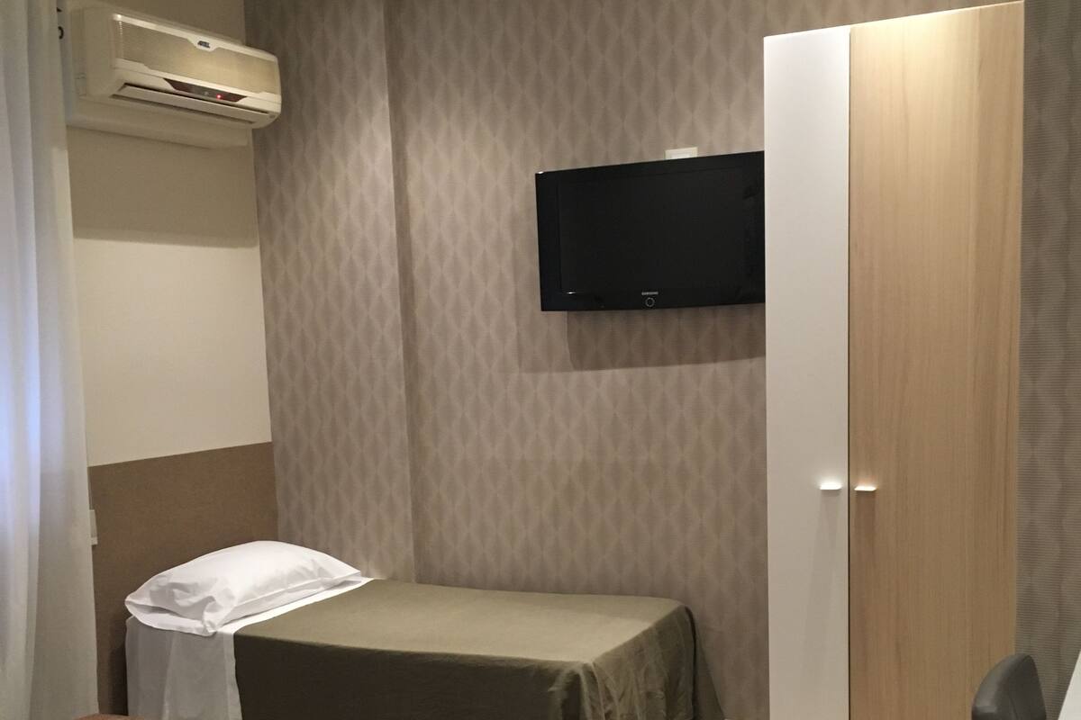 A hotel room with two beds, a TV mounted on the wall, a wardrobe, and an air conditioning unit.