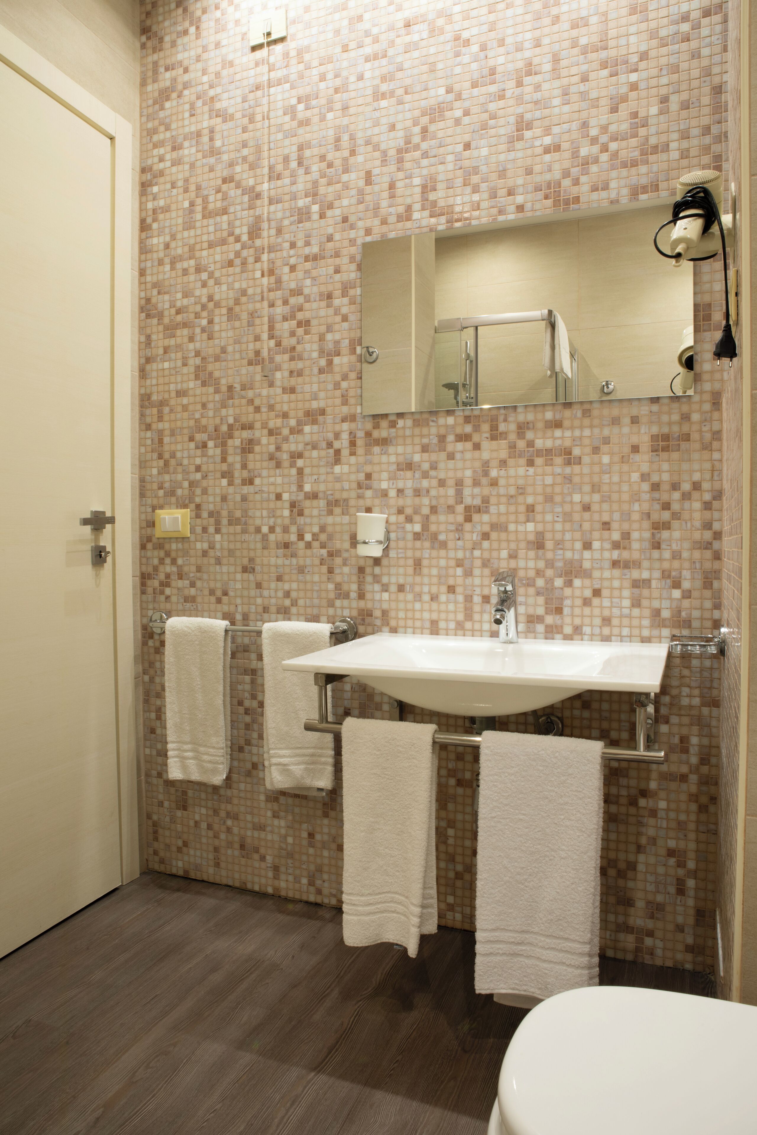 Une salle de bain avec un mur recouvert d’une mosaïque, un lavabo blanc, un miroir et deux serviettes accrochées au mur.