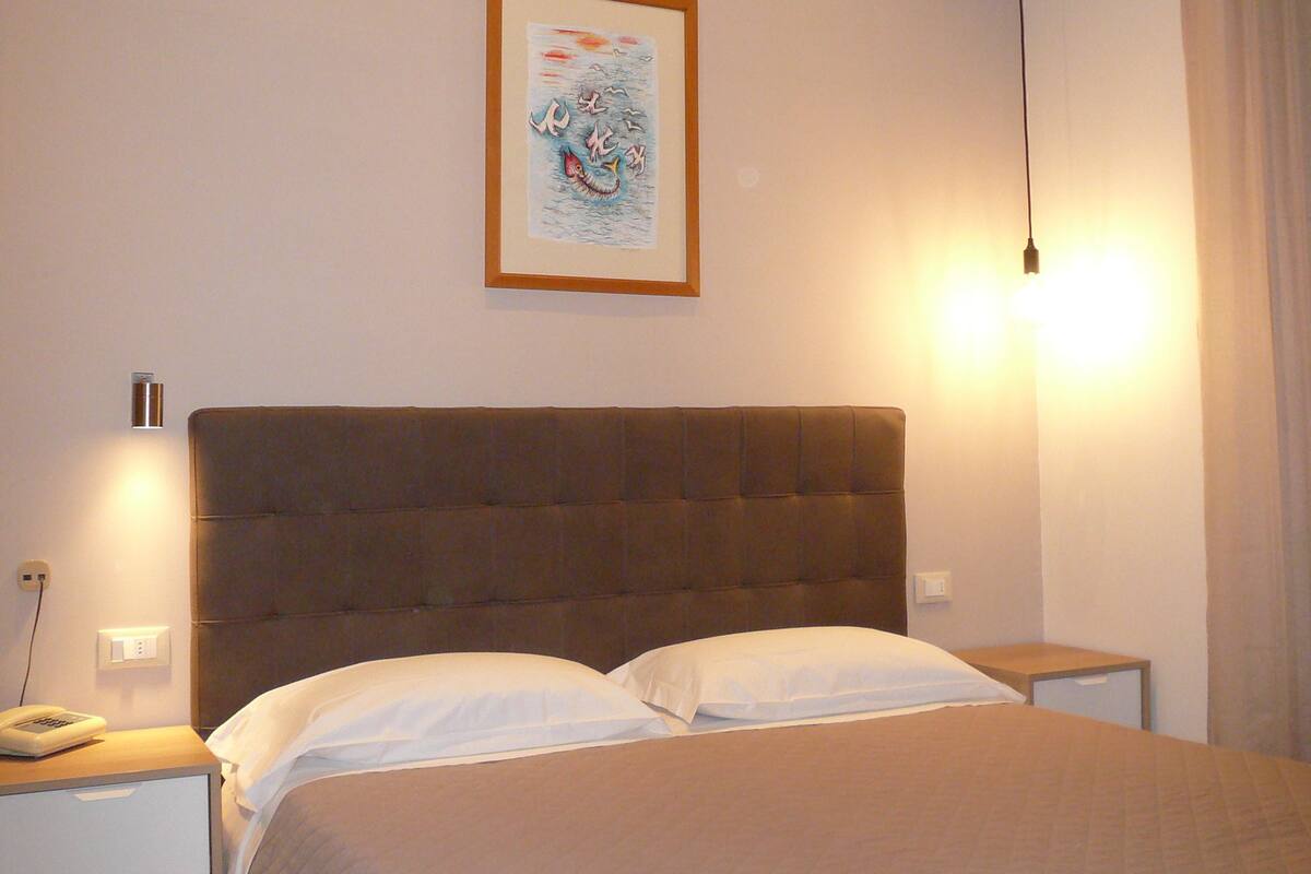 A bedroom with a large bed, a nightstand, a wall-mounted lamp, and a framed picture on the wall.