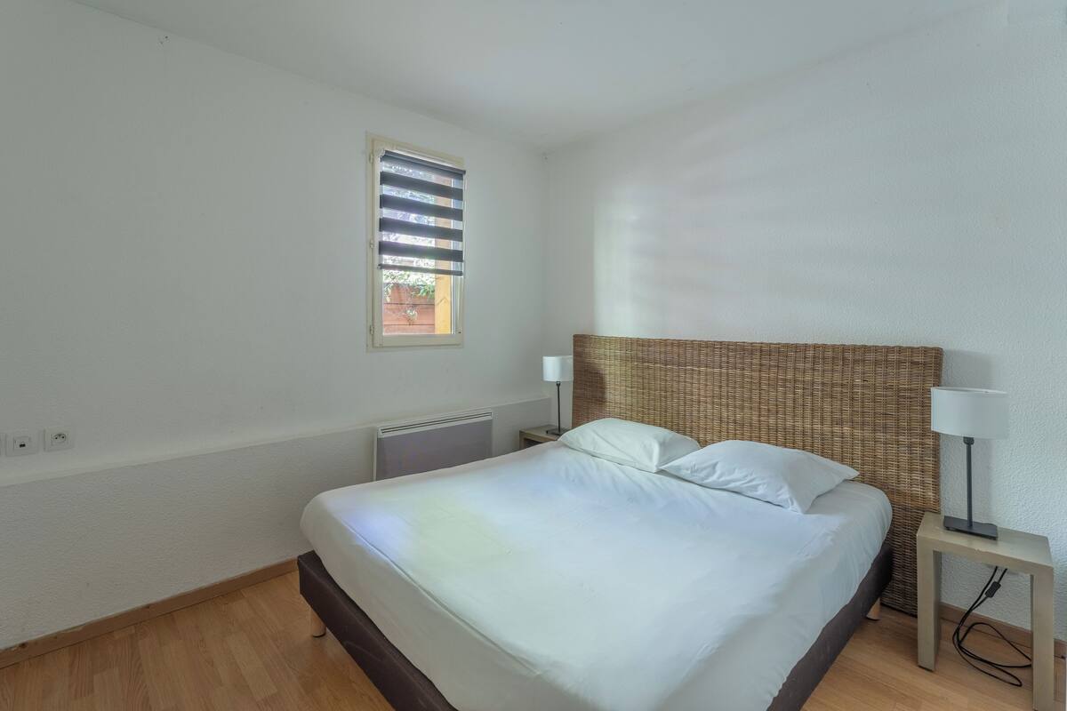 Un dormitorio con cama, ventana, radiador y mesita de noche con lámpara.