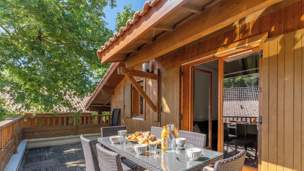 An outdoor wooden deck with a glass-top table and wicker chairs, surrounded by a wooden fence and trees. - Residence Les Rives du Lac - Vacancéole (Lacanau)