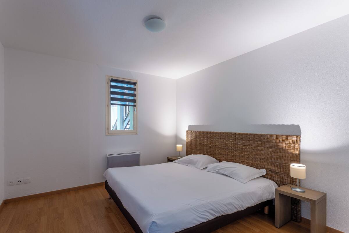 Un dormitorio con cama, mesita de noche, lámpara, ventana y radiador.