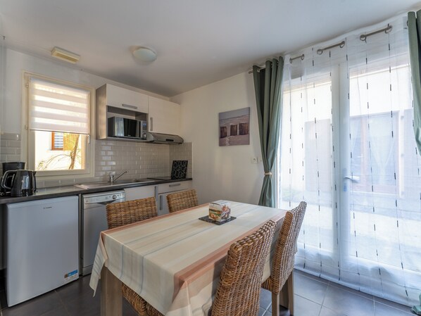 Cocina compacta con microondas, lavamanos y un refrigerador pequeño. Hay una mesa de comedor con sillas y una ventana con cortinas.
