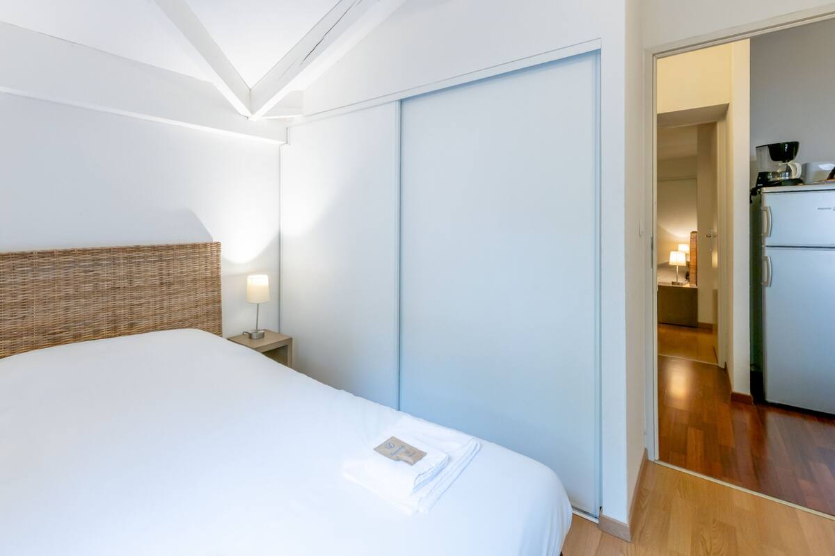 Habitación de hotel con una cama grande, una mesita de noche con lámpara, un armario y una kitchenette al fondo.