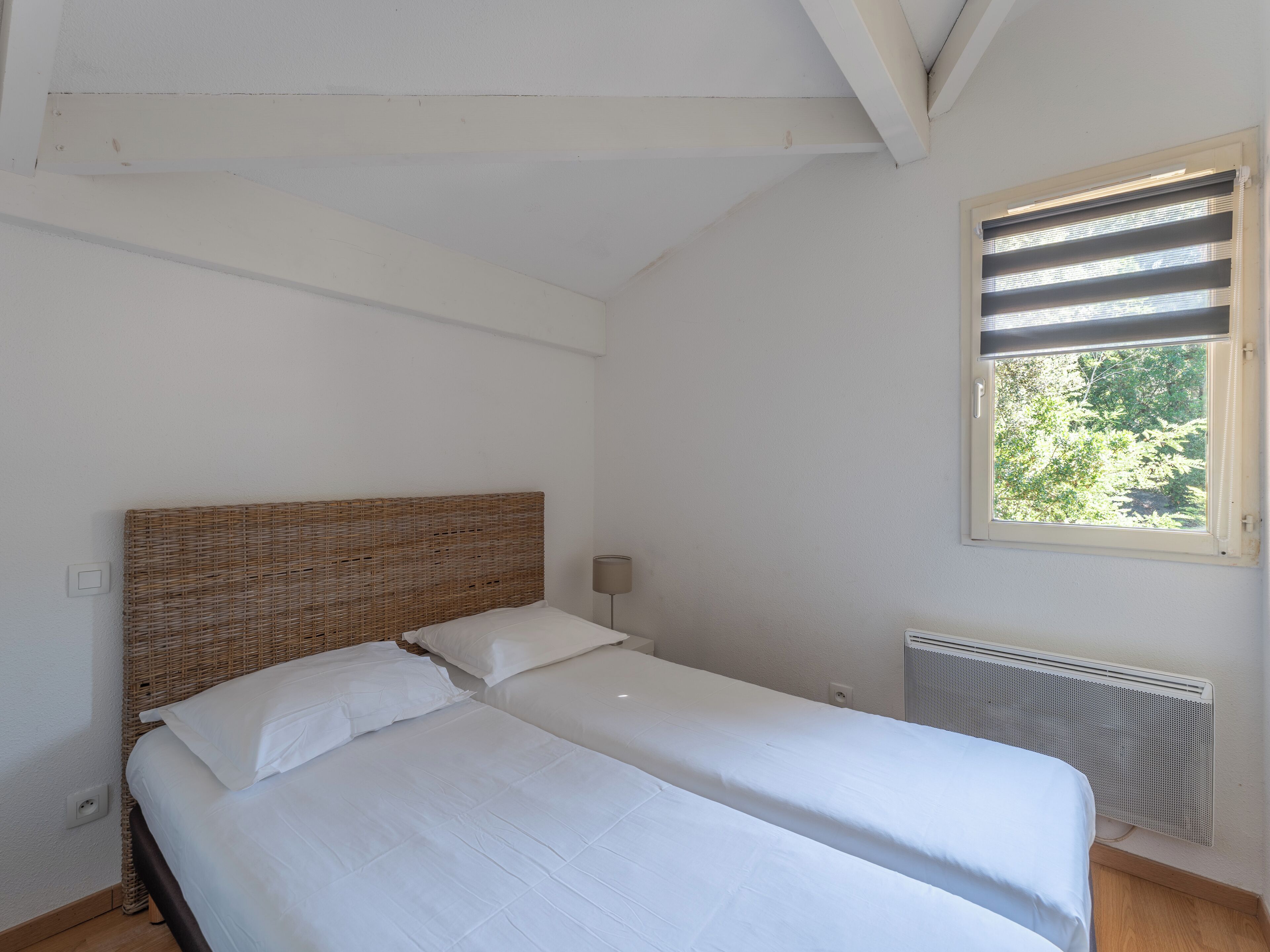 A bedroom with a bed, a window with blinds, a radiator, and a wall switch.