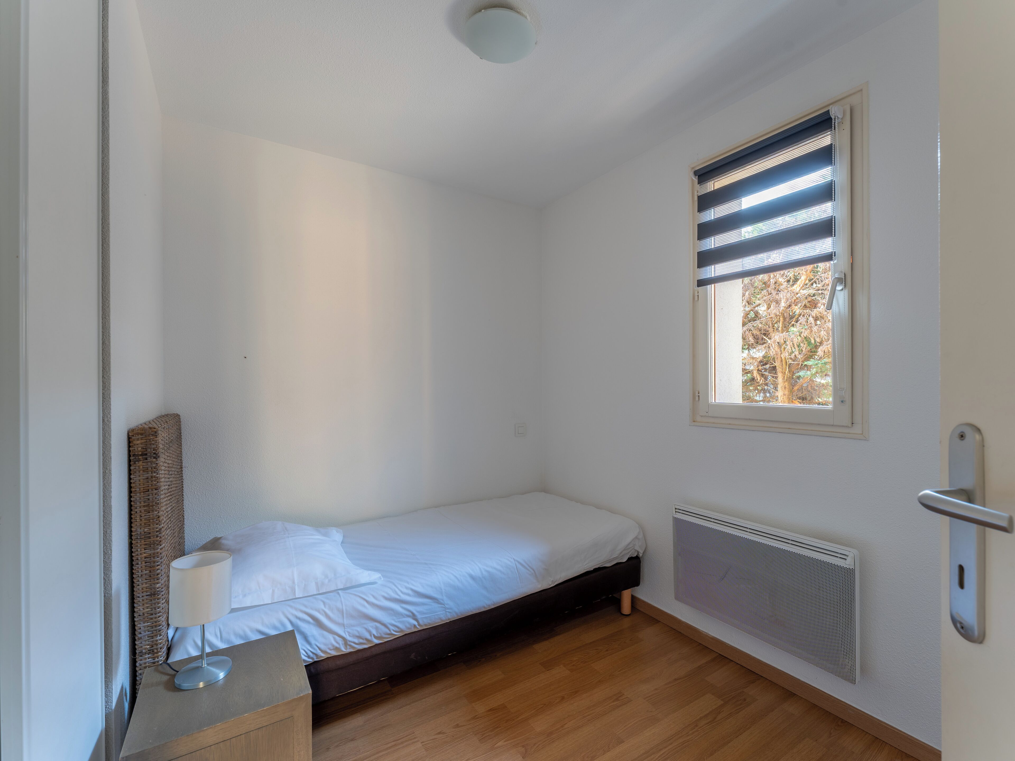 Ein kleines Zimmer mit einem Einzelbett, Holzboden, einem Fenster mit Jalousien, einem Heizkörper und einem Nachttisch mit Lampe.