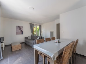 A modern living room with a grey sofa, a dining table with wicker chairs, a flat-screen TV, and a large window with curtains. - Residence Les Rives du Lac - Vacancéole (Lacanau)