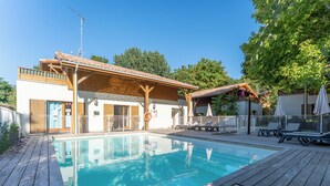 A pool area with wooden deck, lounge chairs, and a building with a tiled roof. - Residence Les Rives du Lac - Vacancéole (Lacanau)