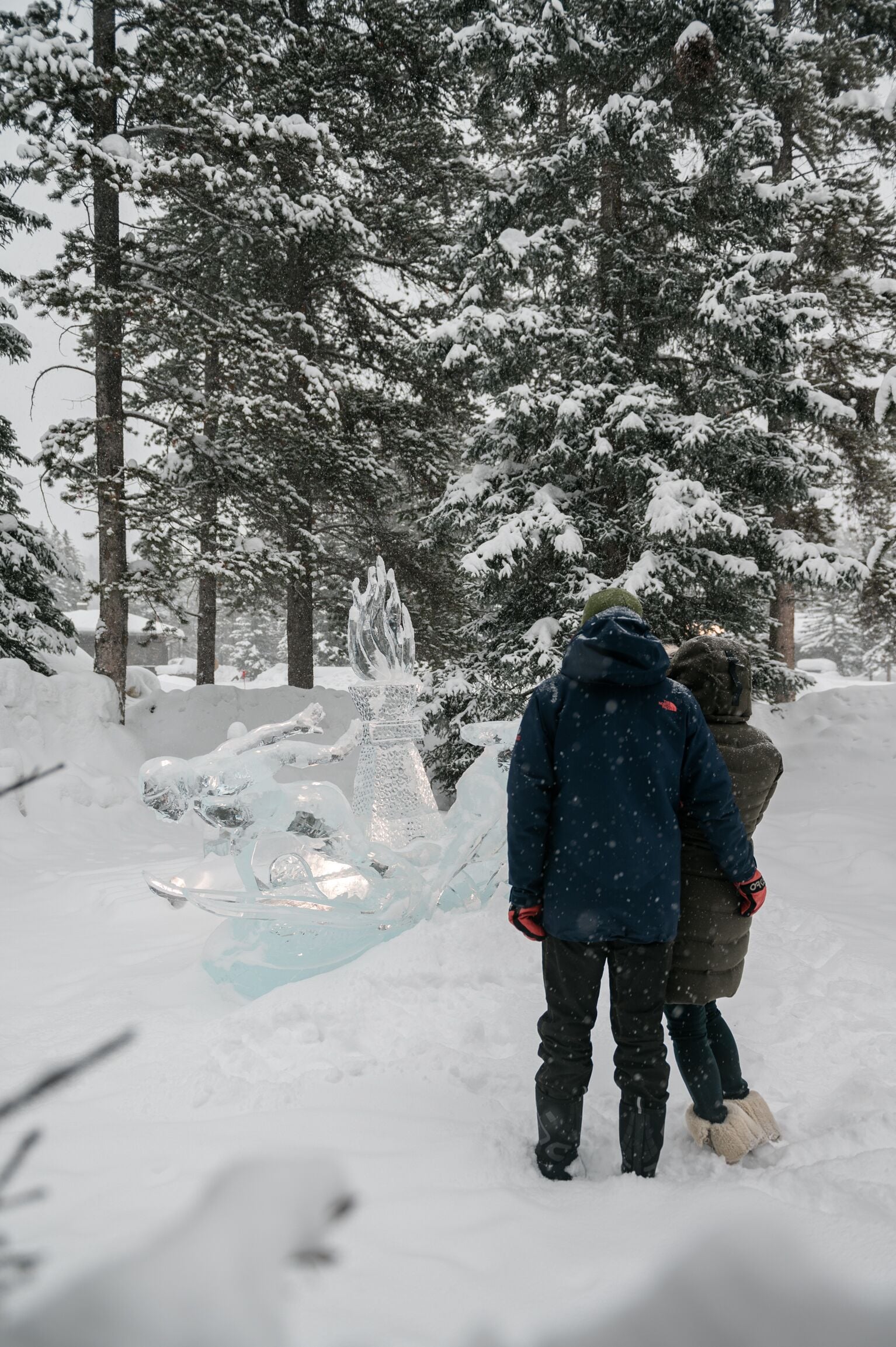 A snowy outdoor scene with a person in a blue jacket and another in a brown jacket standing near a snow sculpture.