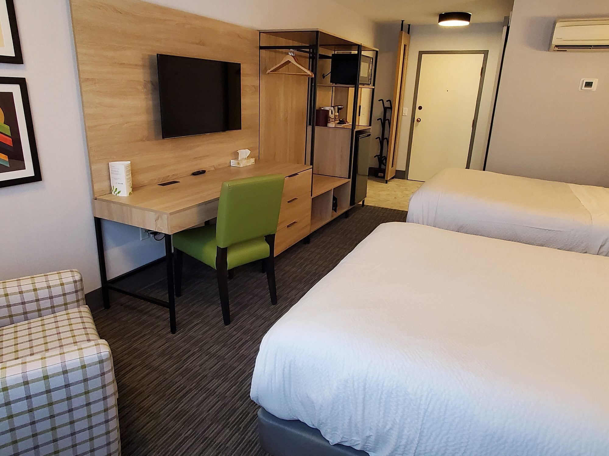 A hotel room with a wooden desk, a green chair, a bed, a wardrobe, and a television.