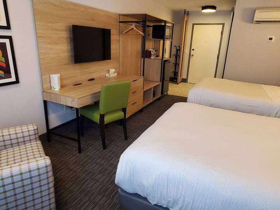 A hotel room with a wooden desk, a green chair, a bed, a wardrobe, and a television.