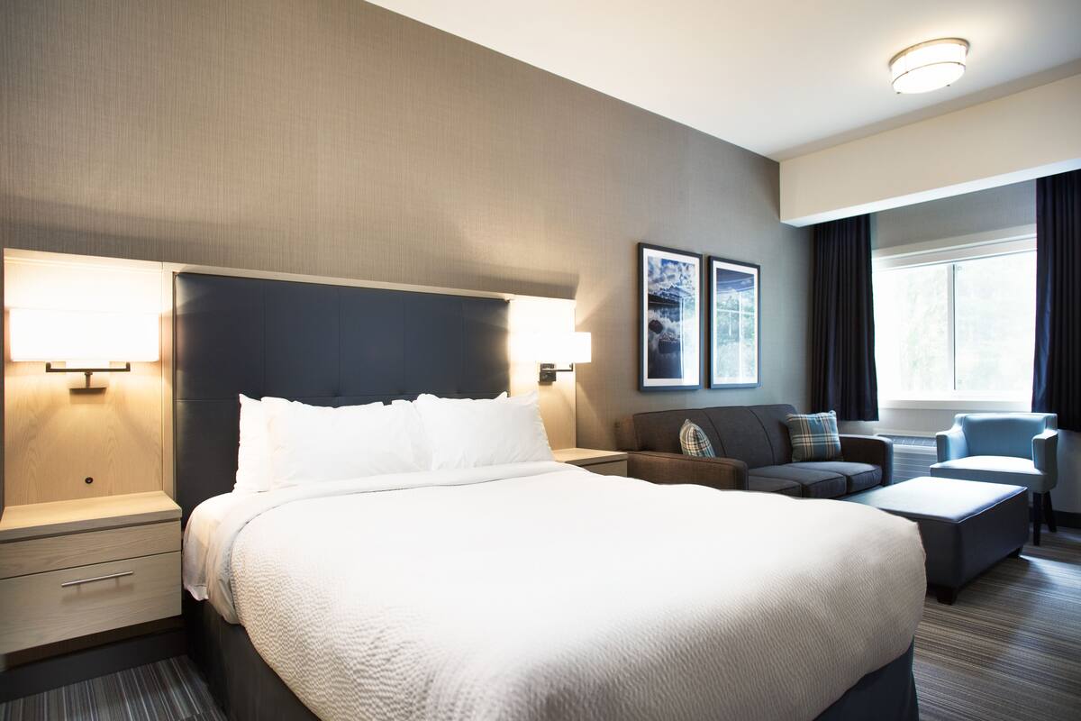 A modern hotel room with a large bed, bedside tables, a sofa, a chair, and a window with curtains.