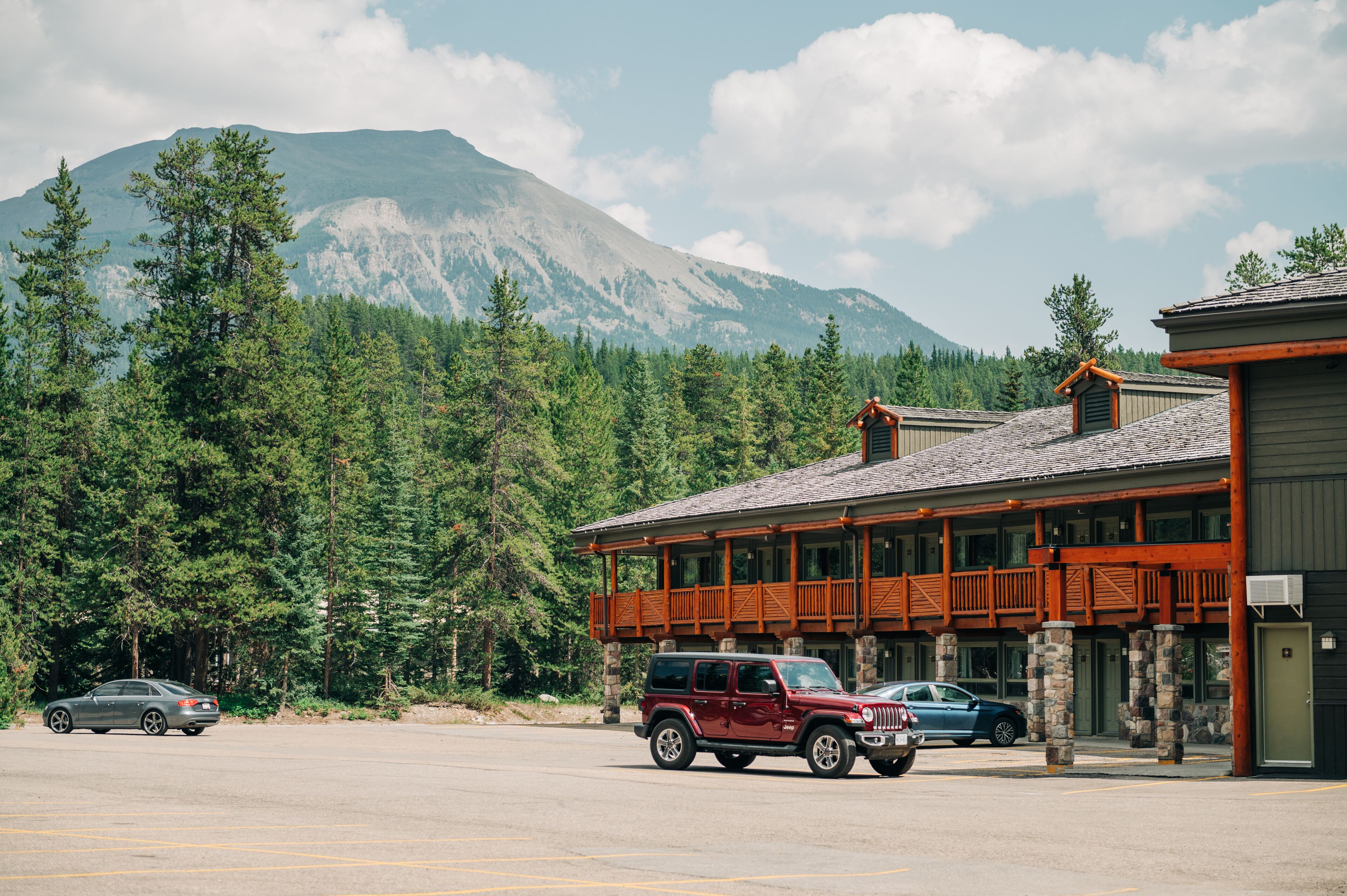 A lodge with a stone facade and wooden balcony, surrounded by forest and mountains.