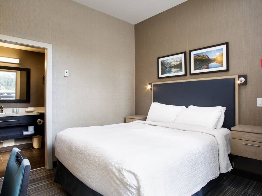 Quarto de hotel com uma cama grande, uma mesa com cadeira, uma cômoda com espelho e duas fotos emolduradas na parede.