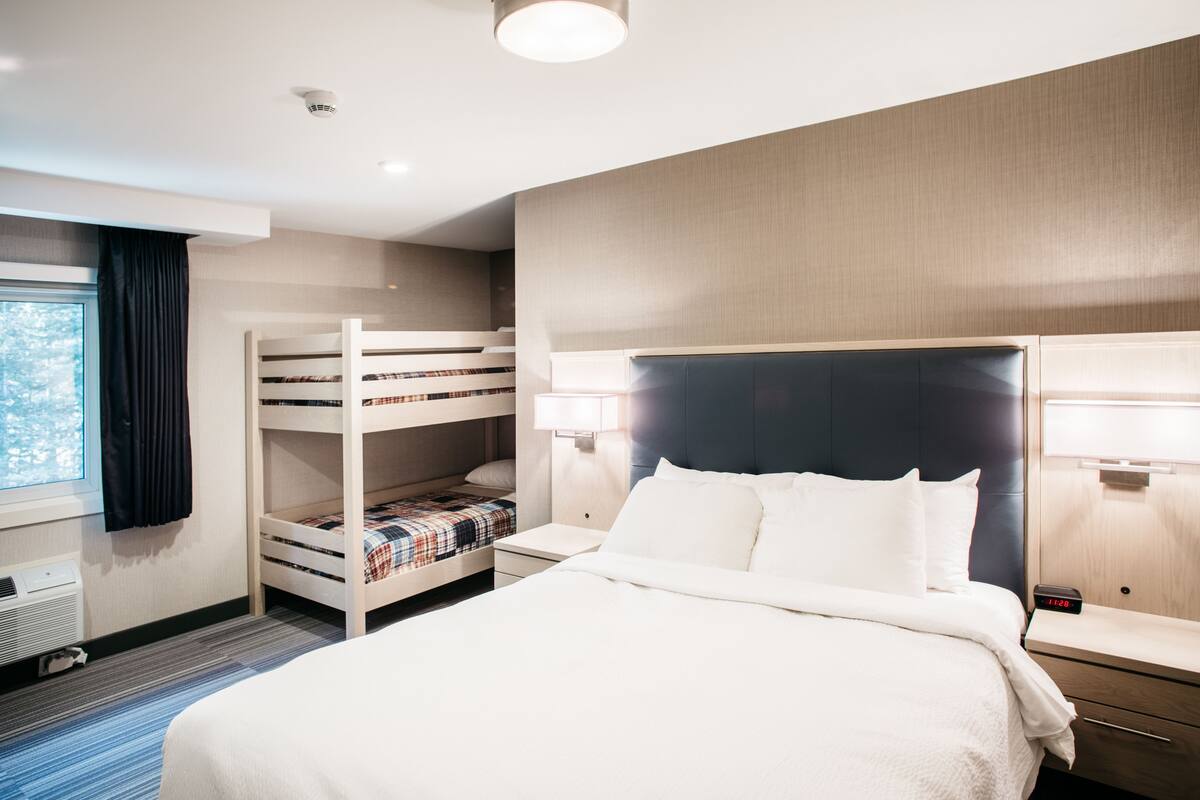 A hotel room with a bunk bed, a single bed, a nightstand, a lamp, and a window with curtains.