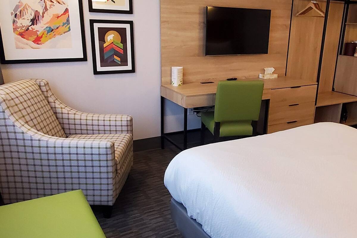 A hotel room with a plaid armchair, a desk with a green chair, a bed, and a TV on the wall.