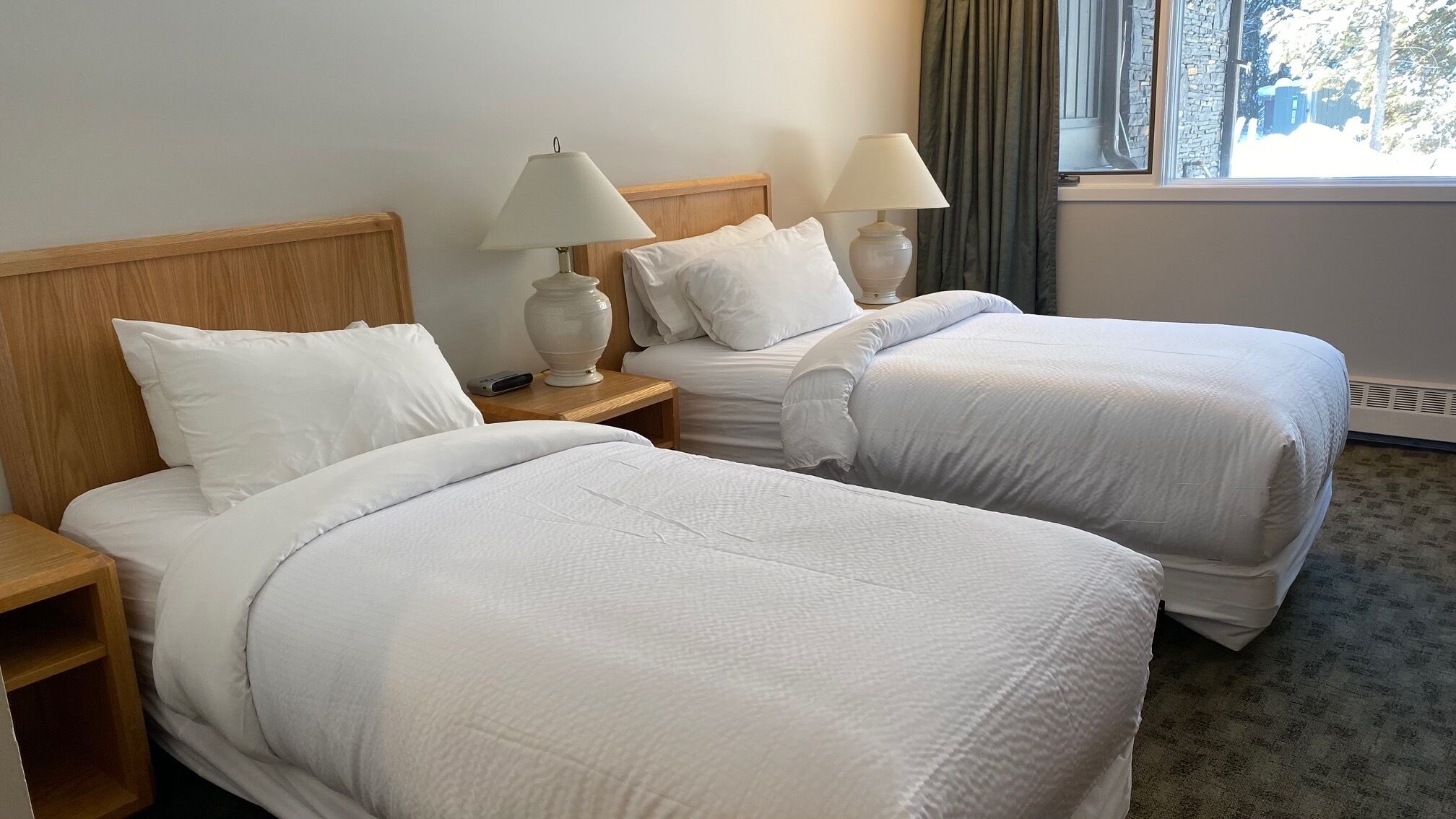 A hotel room with two beds, each with white bedding and pillows, wooden headboards, bedside tables with lamps, and a window with curtains.