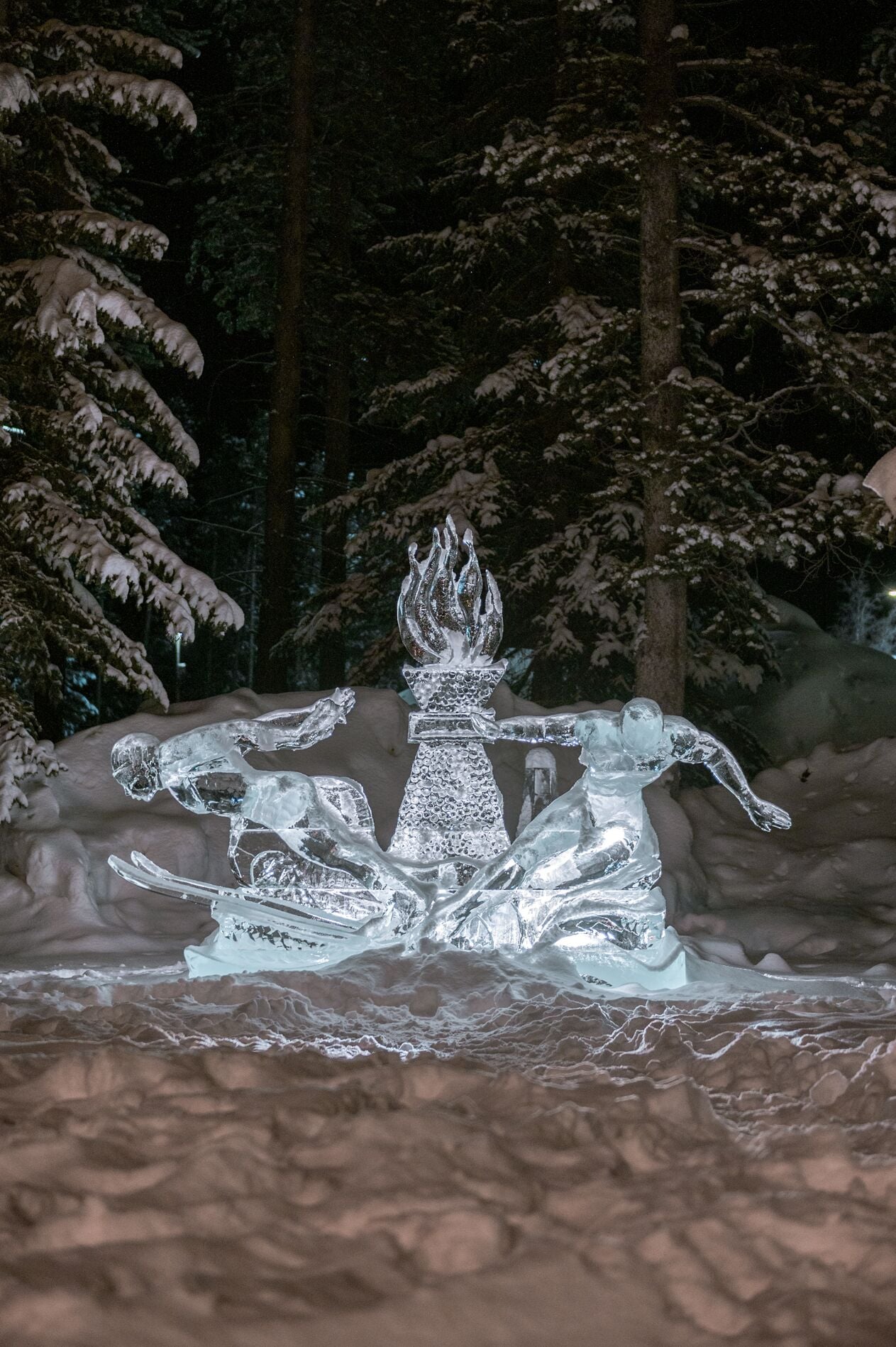 Une sculpture de neige représentant une figure coiffée d’une flamme, entourée d’arbres recouverts de neige.