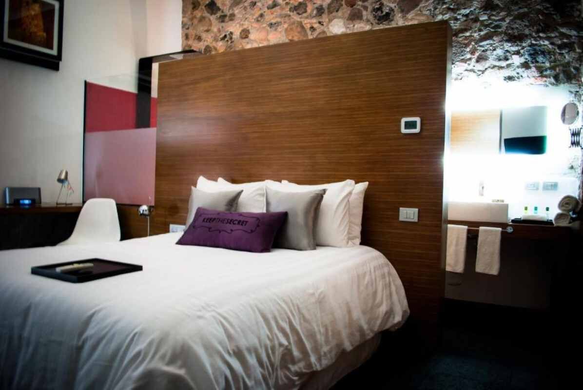 Quarto de hotel com parede de pedra, cabeceira de madeira, cama branca com travesseiro roxo, mesa de cabeceira com abajur e banheiro com pia e porta-toalhas.