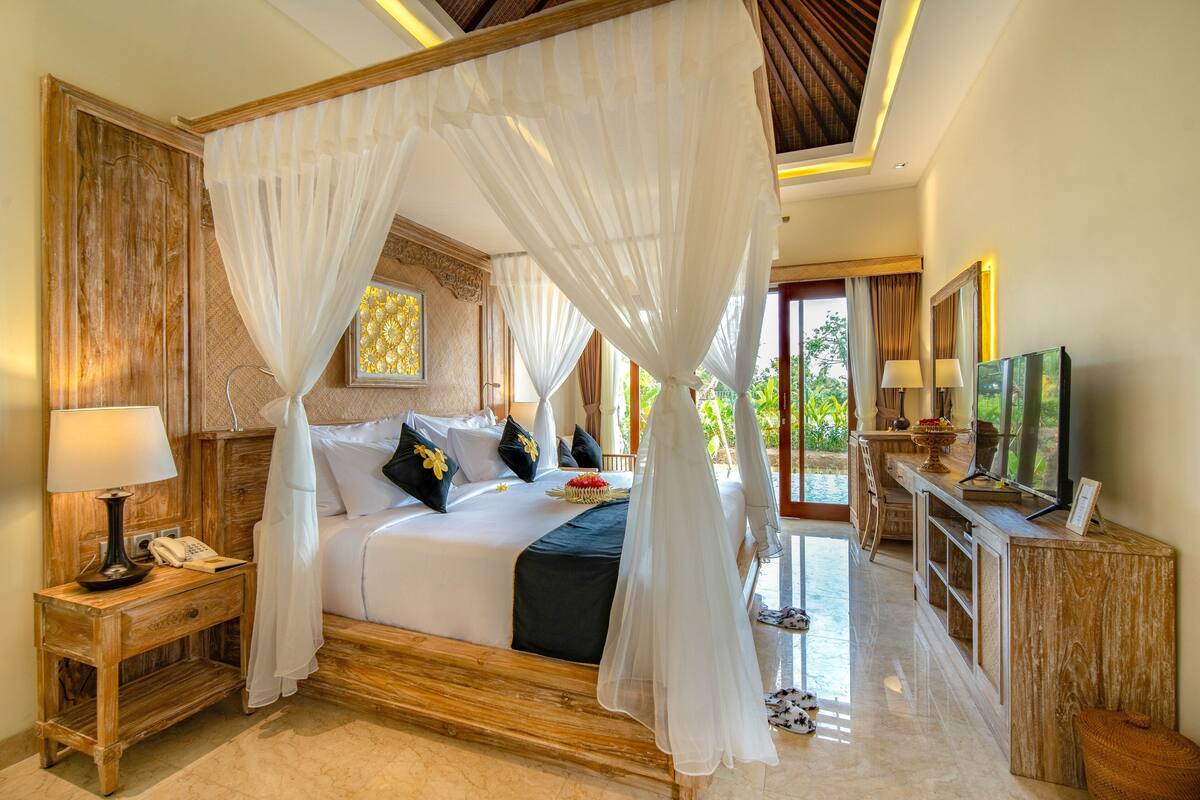 A spacious bedroom with a canopy bed, a wooden nightstand with a lamp, a TV, and a view of the outdoors.