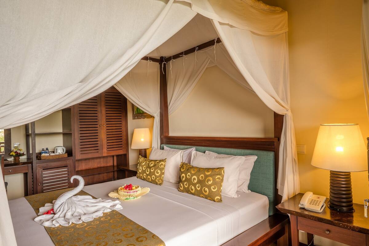 A hotel room with a four-poster bed, a bedside lamp, a telephone, and a decorative swan on the bed.