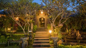 A well-lit garden pathway leading to a building entrance, surrounded by lush greenery and stone sculptures. - Bliss Bungalow (Ubud)