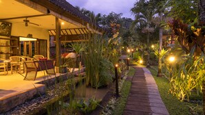 A well-lit outdoor area with a wooden deck, seating, and a pathway lined with plants and lights. - Bliss Bungalow (Ubud)