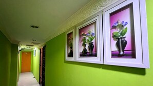 Um corredor com paredes verdes, quadros com flores e uma porta com a inscrição 'KELURAH'.