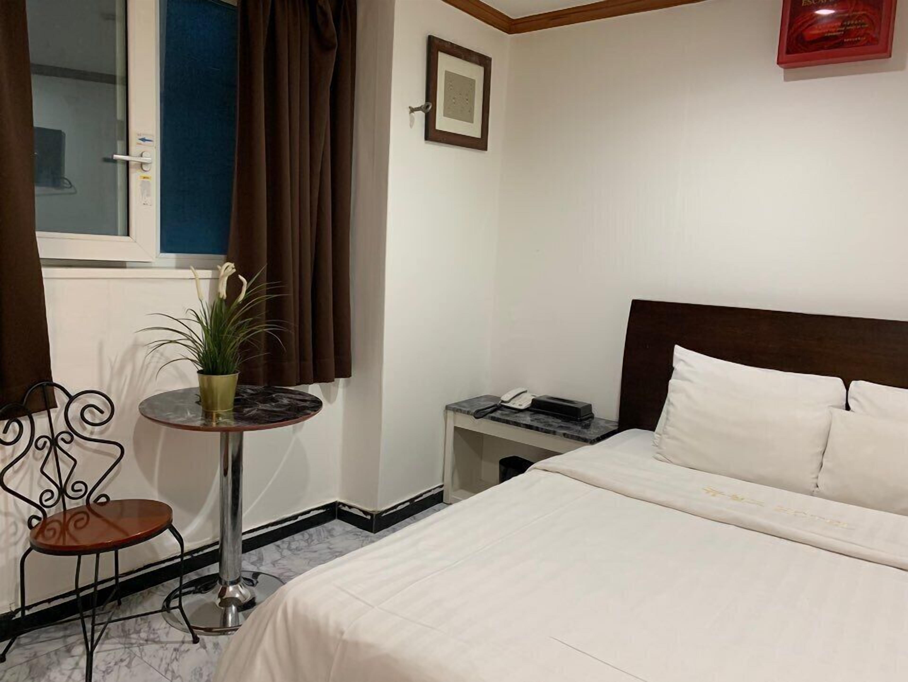 A hotel room with a bed, a small table with a plant, a chair, a nightstand with a phone, and a window with curtains.