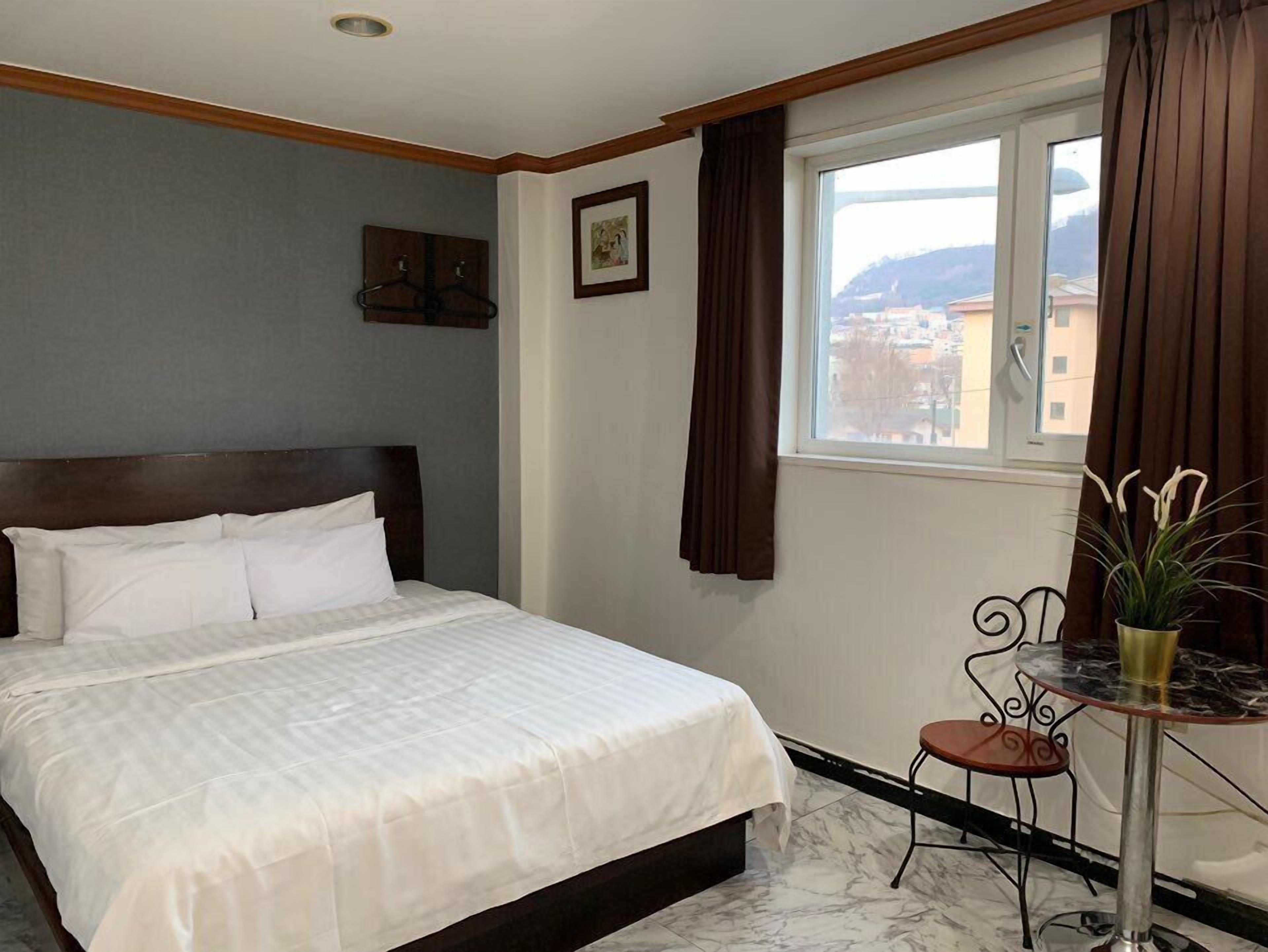 A hotel room with a bed, a small table with a chair, a window with curtains, and a picture on the wall.