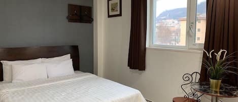 A hotel room with a bed, a small table with a chair, a window with curtains, and a picture on the wall.