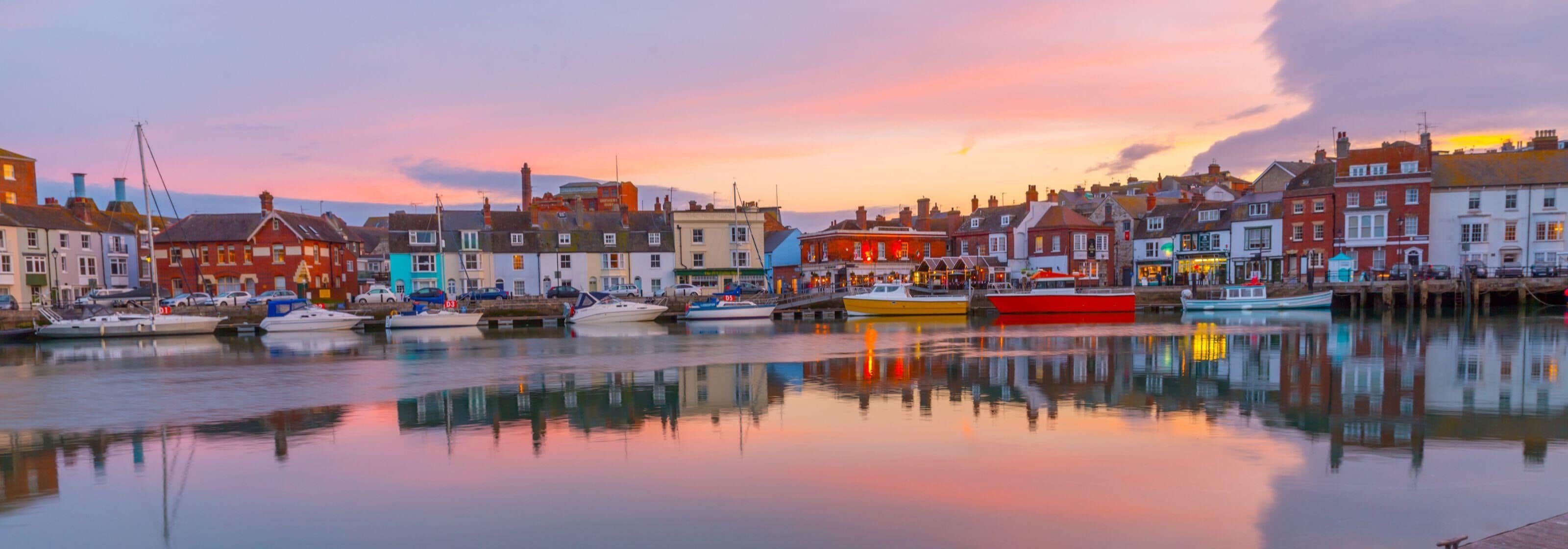 A waterfront town with historic buildings, calm water reflecting the structures, and a sunset sky.