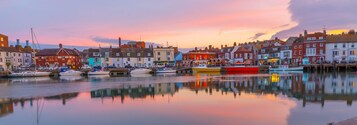 Un pueblo costero con edificios históricos, agua tranquila que refleja las construcciones y un cielo al atardecer.