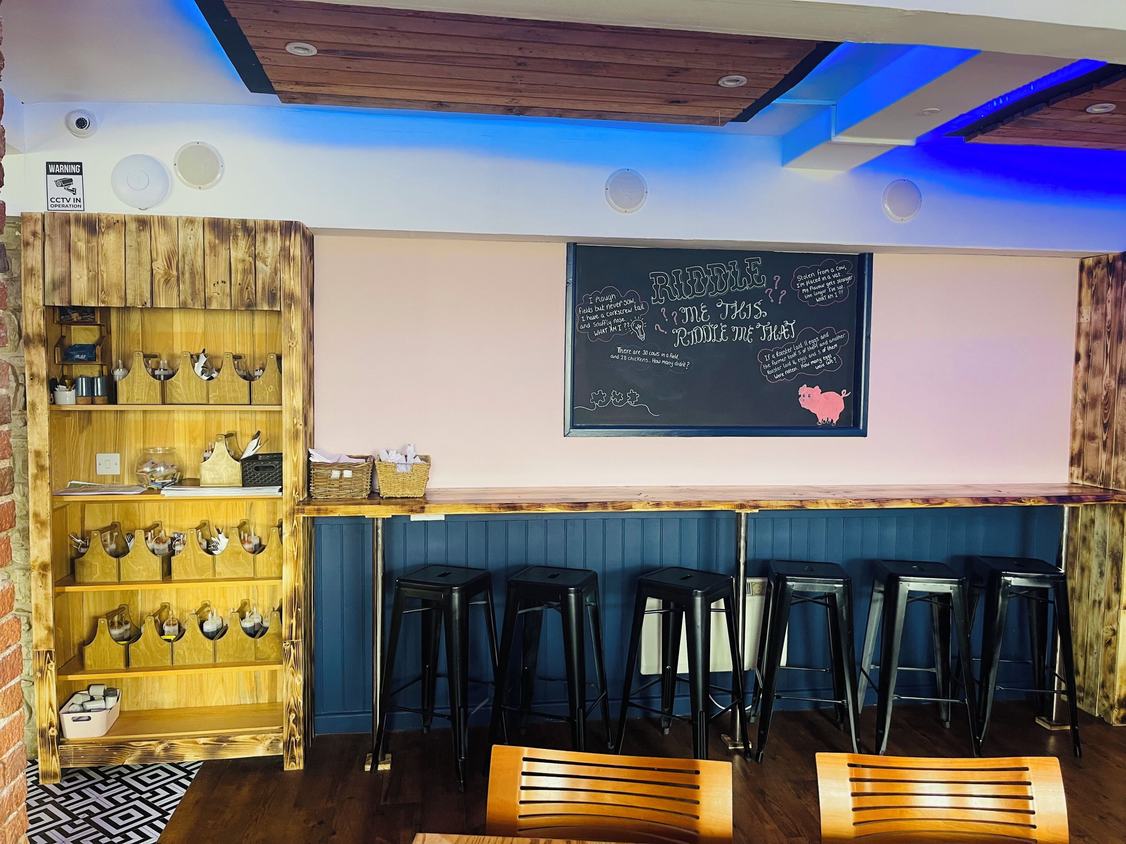 Una zona de bar con estantes de madera, taburetes negros y una pizarra con el menú.